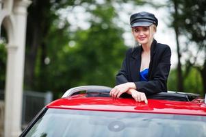 ritratto di bella bionda modello di donna di moda sexy in berretto e in tutto nero con trucco luminoso in tettuccio apribile rosso city car. foto