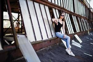 ragazza con occhiali da sole e jeans posati sul tetto di un luogo industriale abbandonato con finestre. foto