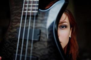 la ragazza punk dai capelli rossi indossa il nero con il basso in un luogo abbandonato. ritratto di musicista donna gotica. primo piano faccia di persona nera con riff di chitarra. foto