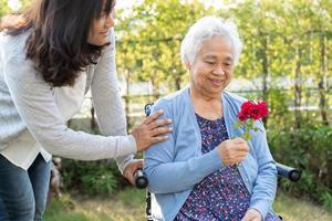 figlia del caregiver abbraccia e aiuta asiatica anziana o anziana signora anziana che tiene una rosa rossa su sedia a rotelle nel parco. foto