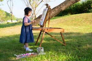 una bambina è in piedi sull'erba e dipinta sulla tela posta su un leggio da disegno foto