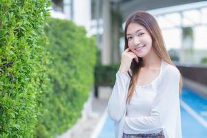 bella donna asiatica con i capelli lunghi in camicia bianca a maniche lunghe si trova fuori dall'edificio mentre le braccia incrociate e guarda la telecamera sorridendo felicemente sull'albero verde come sfondo. foto