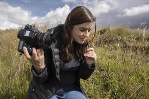 bella fotografa ispanica con la sua macchina fotografica in mano che annusa un fiore giallo in mezzo al campo foto