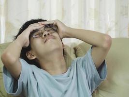 ragazzino asiatico con gli occhiali con la mano sulla testa, mal di testa perché stress. è triste di soffrire di emicrania. foto