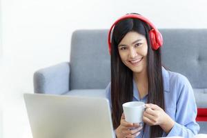 una donna asiatica con una faccia sorridente tiene una tazza di caffè in mano e si diverte con un computer portatile nel soggiorno di casa. foto