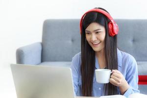 una donna asiatica tiene in mano una tazza di caffè e si diverte con un computer portatile nel soggiorno di casa. foto