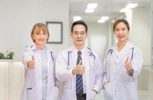 il team di medici sta guardando la fotocamera e sorride mentre è in piedi in ospedale, i medici in piedi come una squadra con i pollici in su foto
