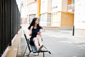 ragazza bruna in abito nero, occhiali da sole seduti su una panchina, ascoltando musica dalle cuffie e posando in una strada della città. foto