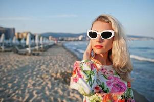 donna bionda che indossa un vestito e occhiali da sole in piedi sulla spiaggia di sabbia in riva al mare godendo della vista del tramonto. foto