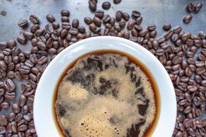 tazza di caffè bianca piena di caffè forte al centro di chicchi di caffè crudo sparsi su un tavolo rustico piatto foto