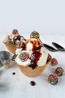 palline di gelato alla vaniglia con caramelle colorate e sciroppo di cioccolato in una ciotola a cono di cialda su sfondo bianco rustico foto