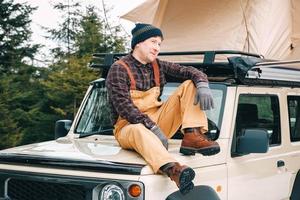 uomo avventuriero seduto sul tetto dell'auto con la tenda montata su di esso per lo stile di vita del campeggio di viaggio gratuito foto