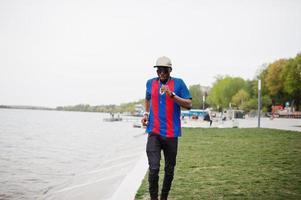 ragazzo afroamericano alla moda che corre contro l'usura del lago al berretto, t-shirt da calcio e occhiali da sole. ritratto di uomo sportivo nero. foto