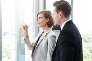 uomo d'affari di successo e donna d'affari che scrivono buone idee su note adesive incollate sulla parete di vetro in una moderna riunione d'ufficio. foto
