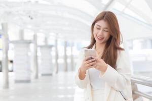 la giovane donna asiatica d'affari in abito bianco sorride e tiene uno smartphone nelle sue mani in una felice giornata di lavoro nella città all'aperto come sfondo. foto