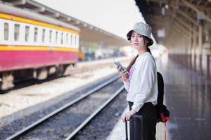 una donna asiatica porta una borsa e uno smartphone in cerca di informazioni di viaggio. in attesa del treno al binario foto