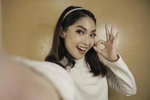 la giovane donna asiatica è selfie con la faccia sorridente felice e il segno giusto vicino alla bocca guarda la telecamera foto