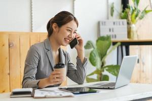 giovane donna d'affari al telefono in ufficio. donna d'affari che manda un sms al telefono e lavora al computer portatile. donna d'affari piuttosto giovane seduta sul posto di lavoro. donna d'affari sorridente. foto