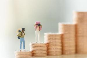 risparmio di viaggio e concetto di planata. due uomini e donne viaggiatori in miniatura figura con zaino in piedi su una pila di monete. foto