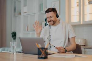 e concetto di apprendimento. felice uomo barbuto studente ha studi in videoconferenza onde online palmo nel display del laptop saluta l'insegnante lavora a distanza usa l'auricolare prende appunti nel diario pone sopra l'interno della casa foto