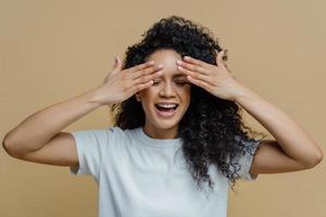 l'inquadratura orizzontale di una donna felice dalla pelle scura copre gli occhi e sorride, si diverte e nasconde il viso, ha i capelli ricci, vestita con una maglietta bianca casual, isolata su sfondo marrone, aspetta la sorpresa. foto