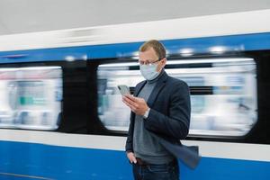 foto di uomo con occhiali e maschera medica protettiva, usa un telefono moderno, viaggia con i mezzi pubblici, si oppone al treno in movimento sulla piattaforma, ottiene informazioni online sul nuovo virus covid-19