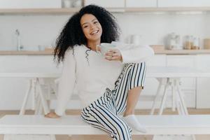 bella giovane donna con taglio di capelli afro, posa su una panca bianca al coperto, vestita con abiti eleganti, gode di una bevanda aromatica, fa colazione a casa, posa contro l'interno sfocato della cucina foto