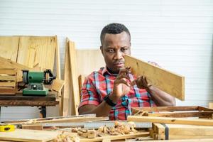 Giovane falegname afroamericano uomo che guarda e sceglie il legno e usa la carta vetrata per strofinare la tavola di legno al tavolo dell'officina nella fabbrica di legno del falegname foto