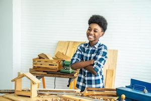 sorridente ragazzo afro-americano falegname in piedi con le braccia incrociate che mostra fiducia dopo aver completato la lavorazione del legno che ha aiutato mio padre a completare foto