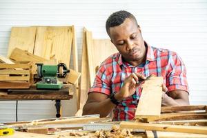 Giovane falegname afroamericano uomo che guarda e sceglie il legno e usa la carta vetrata per strofinare la tavola di legno al tavolo dell'officina nella fabbrica di legno del falegname foto