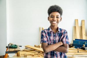 sorridente ragazzo afro-americano falegname in piedi con le braccia incrociate che mostra fiducia dopo aver completato la lavorazione del legno che ha aiutato mio padre a completare foto