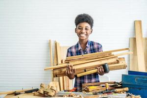 sorridente ragazzo afro-americano falegname in piedi tenere diversi pezzi di legno per aiutare il padre a prepararsi a fare artigianato di lavorazione del legno foto