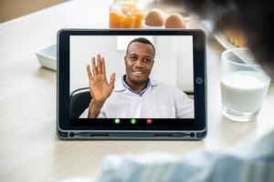 primo piano del padre afroamericano che saluta mentre comunica tramite videochiamata con sua figlia che lo chiama mentre beve il latte mattutino da casa. foto