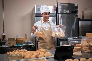 chef maschio asiatico senior in grembiule, blogger di piccole imprese in diretta streaming tramite tecnologia di applicazione mobile, promozione online di vendita di prodotti da forno, pane fresco, pasticcini e cucina commerciale in cucina. foto