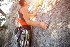 lo scalatore in maglietta rossa si arrampica su una roccia grigia. una mano forte afferrò il comando, messa a fuoco selettiva. forza e resistenza, attrezzatura da arrampicata corda, imbracatura, gesso, sacca portamagnesite, moschettoni, bretelle, rinvii foto