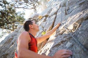 lo scalatore in maglietta rossa si arrampica su una roccia grigia. una mano forte afferrò il comando, messa a fuoco selettiva. forza e resistenza, attrezzatura da arrampicata corda, imbracatura, gesso, sacca portamagnesite, moschettoni, bretelle, rinvii foto