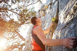 lo scalatore in maglietta rossa si arrampica su una roccia grigia. una mano forte afferrò il comando, messa a fuoco selettiva. forza e resistenza, attrezzatura da arrampicata corda, imbracatura, gesso, sacca portamagnesite, moschettoni, bretelle, rinvii foto