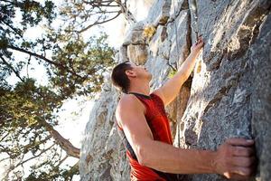 lo scalatore in maglietta rossa si arrampica su una roccia grigia. una mano forte afferrò il comando, messa a fuoco selettiva. forza e resistenza, attrezzatura da arrampicata corda, imbracatura, gesso, sacca portamagnesite, moschettoni, bretelle, rinvii foto