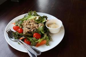 insalata di tonno e condimento per insalata in una tazzina in un piatto rotondo bianco su un tavolo marrone scuro. foto