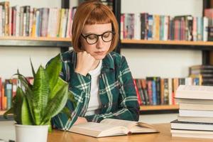 giovane donna rossa con gli occhiali legge il libro in biblioteca foto