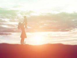 una silhouette di una bambina felice le braccia della sua amorevole madre per un abbraccio, davanti al tramonto nel cielo in una giornata estiva. foto