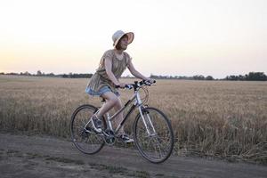 giovane donna con cappello giro in bicicletta nei campi di grano estivi foto