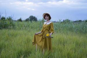 giovane donna rossa con le lentiggini in abito vintage fatto a mano cammina nei campi con fiori foto