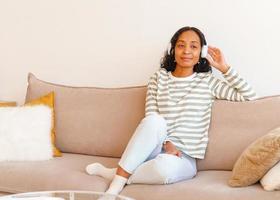 sorridente femmina afro-americana che ascolta musica in cuffia mentre è seduto sul divano foto