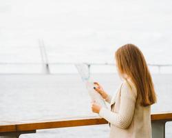 giovane donna in piedi sul lungomare della grande città e guardando la guida, un turista in porto in cerca di attrazioni e in attesa del traghetto foto