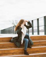 bella donna con i capelli lunghi che parla al telefono e cerca qualcosa in lontananza. autunno o inverno, ragazza in giacca esterna seduta su gradini di legno foto