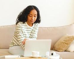 femmina afro-americana seduta sul divano e che utilizza un dispositivo digitale per lavoro e studio foto