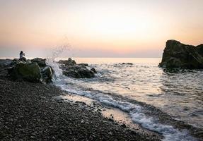 la donna da lontano si siede sulla roccia e scatta una foto del mare con le onde che si infrangono sullo sfondo. vacanza sul Mar Nero e spiaggia rocciosa.