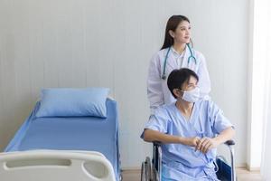 donna medico asiatica che parla con un paziente uomo che indossa una maschera sul suo sintomo di salute mentre si siede su una sedia a rotelle in ospedale. foto