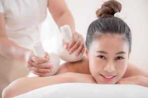 una bella donna asiatica è rilassata e rilassante, la massaggiatrice usa un impacco caldo alle erbe in un negozio di terme foto